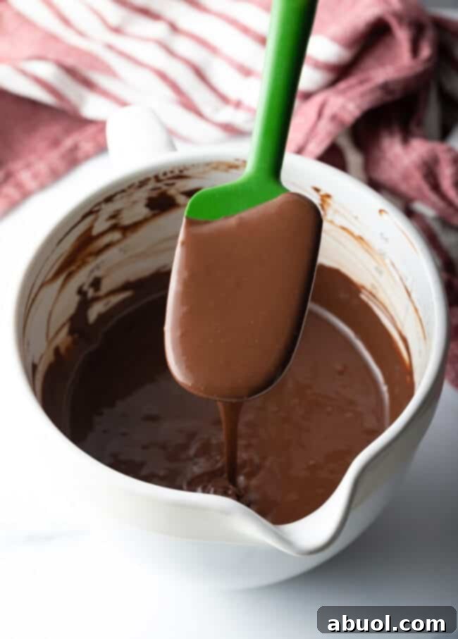 Green spatula held over a white bowl with homemade hot fudge sauce.