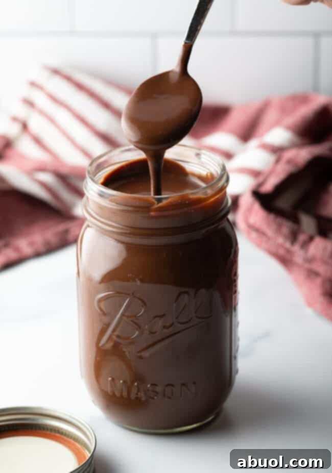 Metal spoon pouring homemade chocolate fudge sauce into a glass mason jar.
