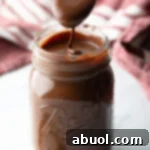 Metal spoon pouring homemade chocolate fudge sauce into a glass mason jar.
