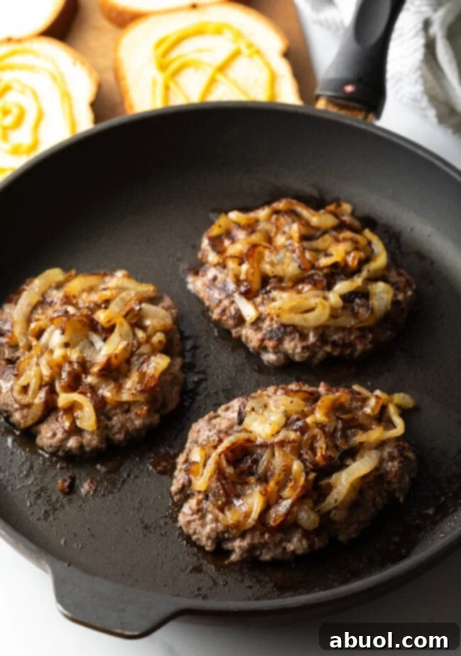 Three oval burger patties cooking in a skillet, each topped with a mound of caramelized onions.