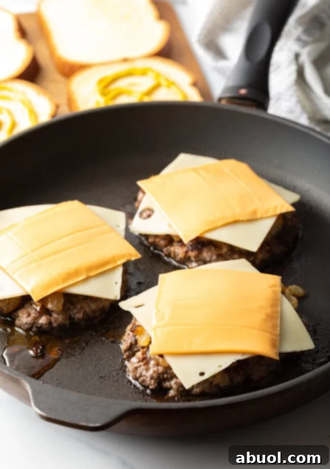 Three burger patties in a skillet, each with a slice of Swiss cheese and a slice of American cheese melting on top.