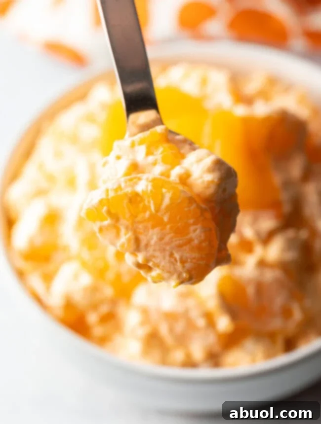 A spoon presents a generous scoop of the creamy Creamsicle Fluff Salad to the camera, highlighting its enticing texture and delightful orange hue.