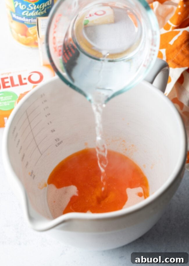 A stream of boiling water being poured from a glass measuring cup into a white mixing bowl, ready to dissolve the gelatin mix.