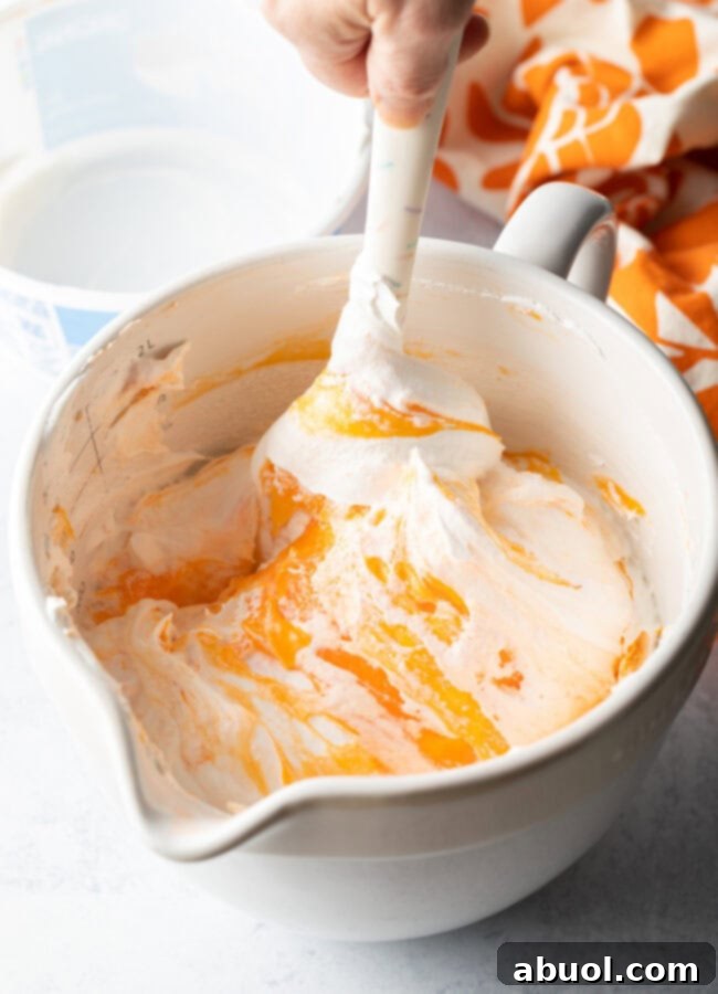 A white spatula gently mixes the orange fluff mixture in a white bowl, creating a light and airy texture.