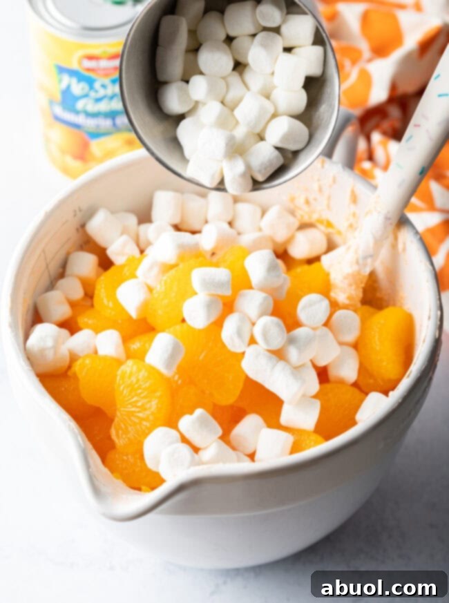 Mini marshmallows are being added to a white mixing bowl containing the fluffy orange mixture and drained mandarin oranges, ready to be gently folded in.