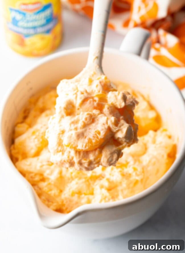 A spoon lifts a generous scoop of the creamy, orange-hued Creamsicle Fluff Salad towards the camera, showcasing its delightful texture and orange pieces.