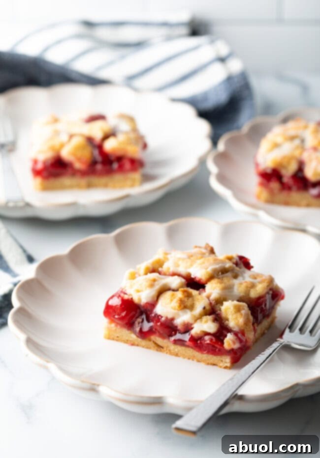 Three perfectly cut pieces of cherry pie bars, each presented on a white scalloped plate, highlighting their appetizing appearance and readiness to be served.