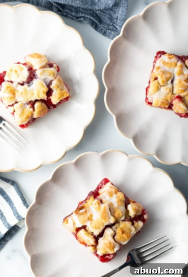 A top-down view of three individual pieces of cherry pie bars, each neatly placed on a white scalloped plate, showcasing their inviting red filling and glazed topping.