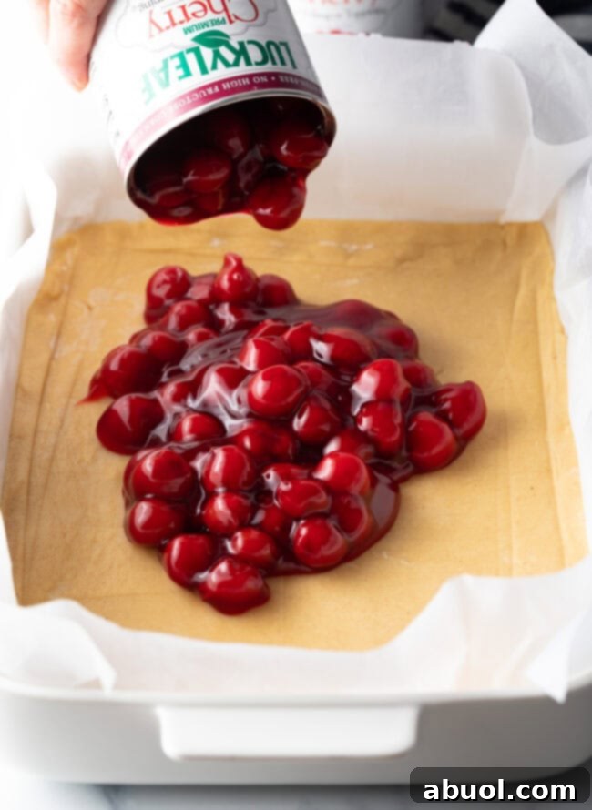 An open can of vibrant red cherry pie filling being carefully poured into a rectangular baking dish, already lined with the prepared dough crust.