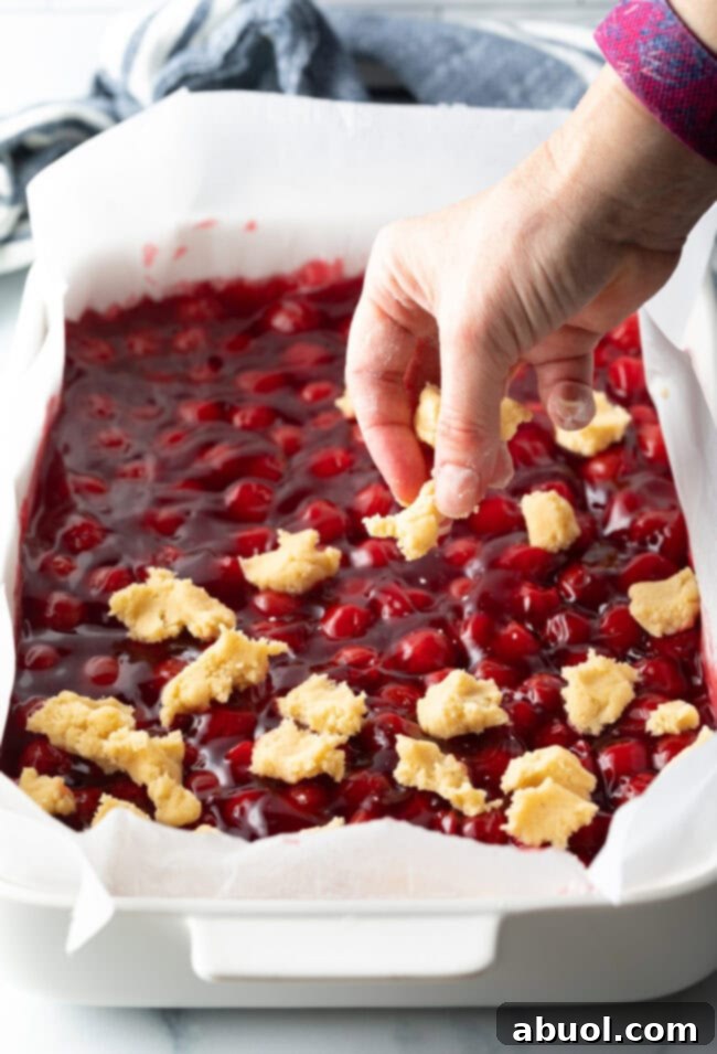 A hand delicately adding small, raw dough pieces, pinched off from the remaining dough, over the surface of the cherry pie filling in the baking dish.