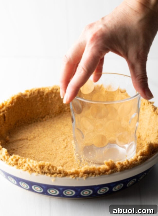 Hand with clear glass, pressing graham cracker pieces into a firm pie crust.