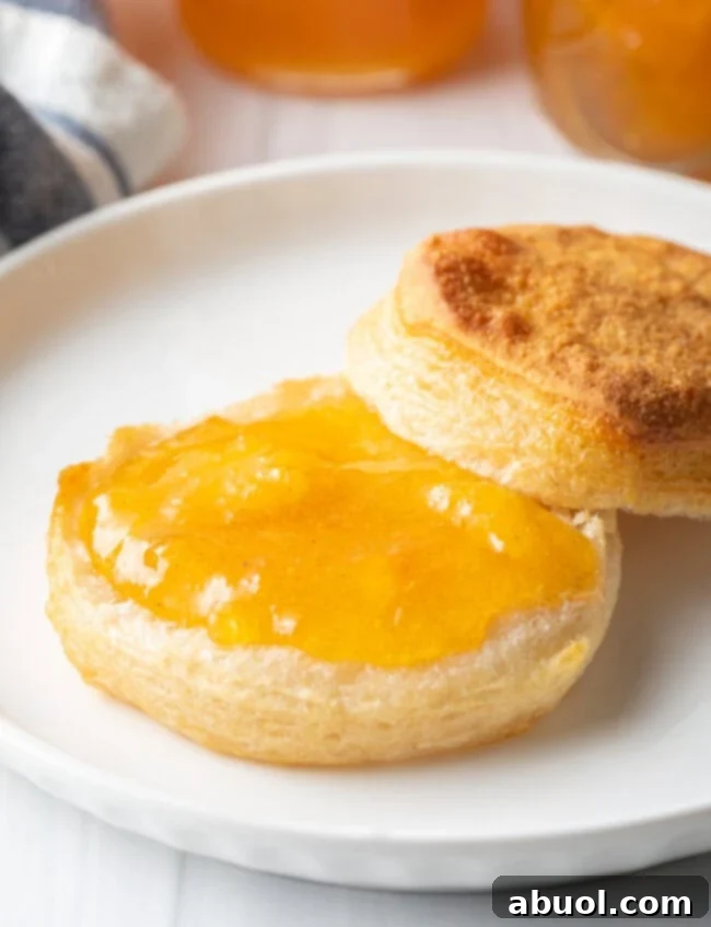 A perfectly split, warm buttermilk biscuit resting on a white plate, with the bottom half generously smeared with glistening homemade peach jam, inviting a delicious bite.