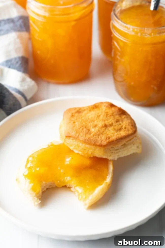 A delicious buttermilk biscuit, perfectly split in half and placed on a pristine white plate. The bottom half is generously smeared with glistening homemade peach jam, and a delightful bite has already been taken, revealing its tender interior.