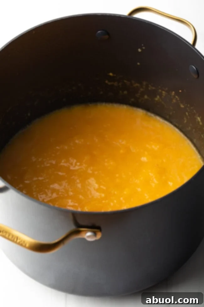 A large black cooking pot filled with bubbling, vibrant cooked peach preserves on a stovetop, indicating active boiling.