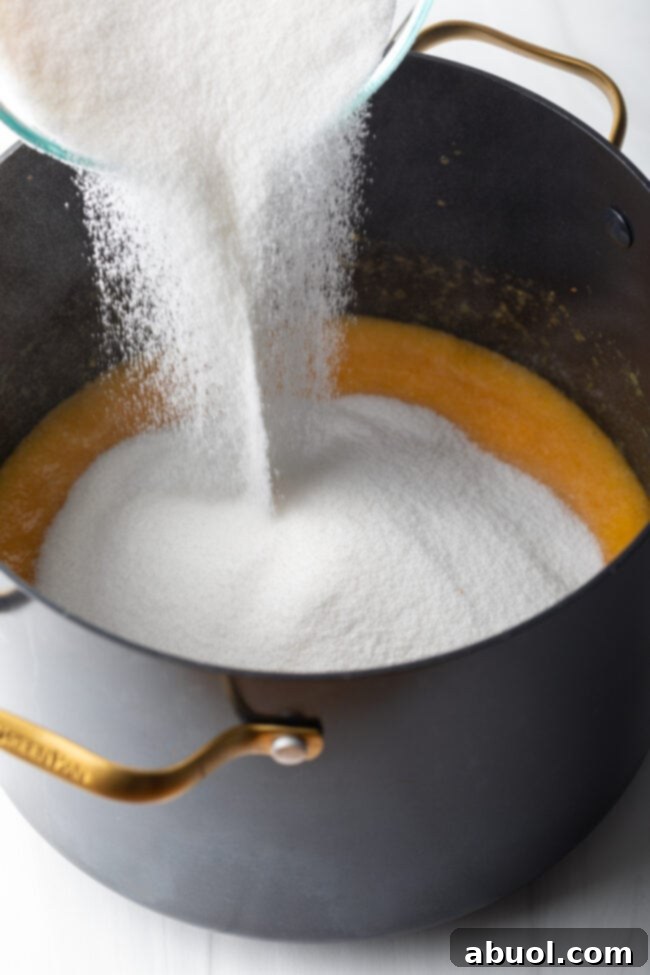A generous amount of granulated sugar being poured into the hot, simmering peach mixture in the pot, crucial for setting and flavor.