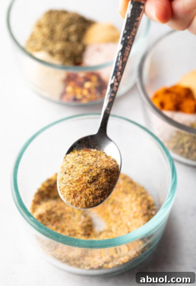 A metal spoon delicately scoops a fresh blend of homemade seasoning from a glass bowl, showcasing its fine texture.