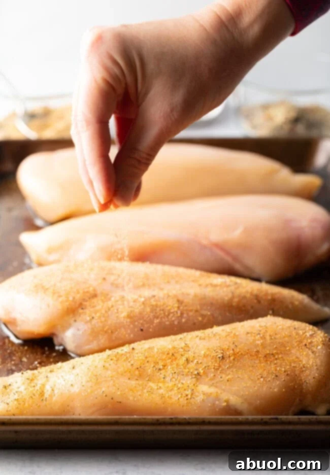 A hand gracefully sprinkles a generous amount of homemade seasoning over raw chicken pieces arranged on a metal baking sheet.