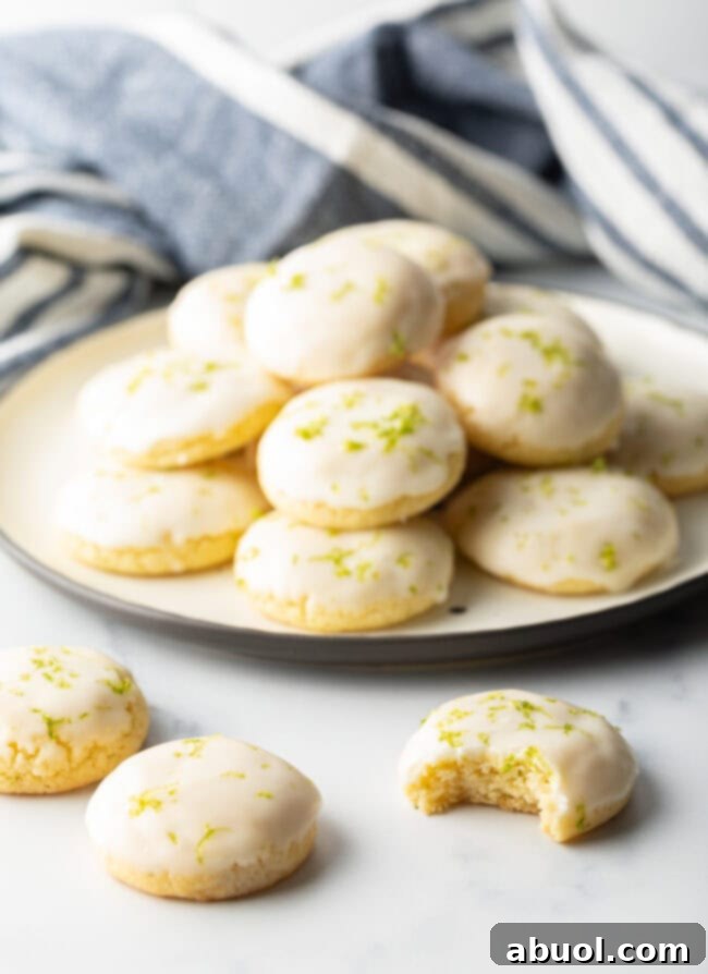 In the background, a plate overflows with perfectly glazed key lime cookies. In the foreground, a few cookies rest on the table, one with a delectable bite taken out, revealing its soft interior.