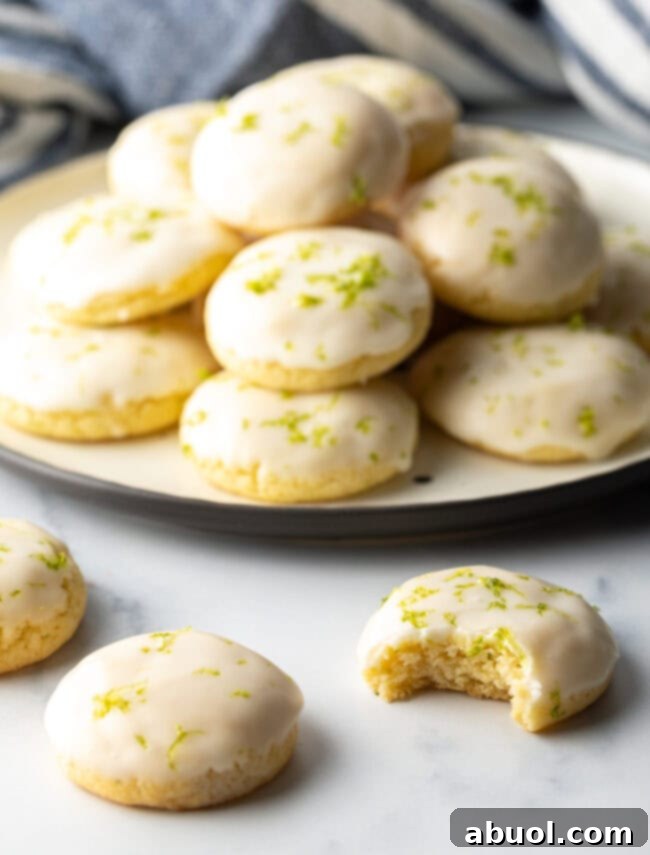 In the background, a plate overflows with perfectly glazed key lime cookies. In the foreground, a few cookies rest on the table, one with a delectable bite taken out, revealing its soft interior.