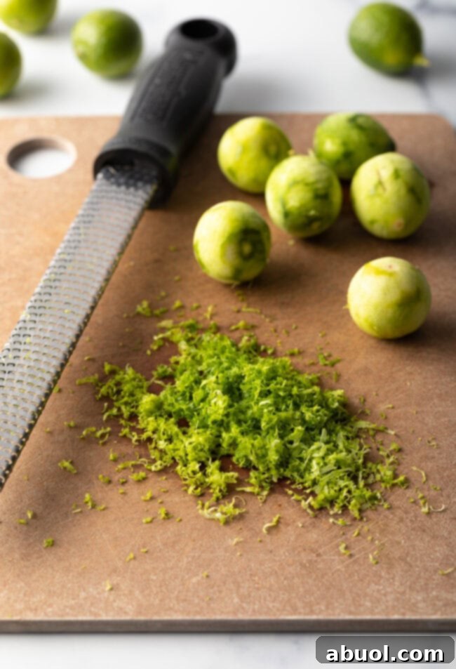 A wooden cutting board displaying several small, vibrant key limes, alongside a zester, and a neat pile of aromatic lime zest, ready for baking.
