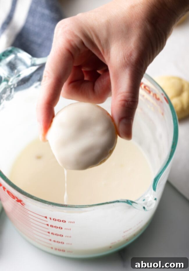 A hand gently holds a single, freshly baked cookie directly above a bowl of glossy glaze, with the cookie's top already coated in the vibrant key lime mixture.