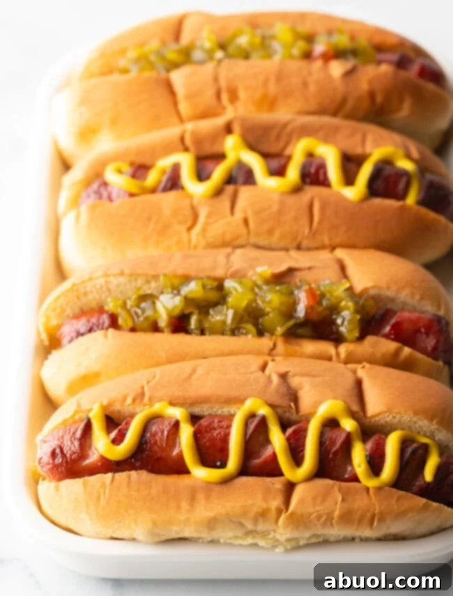 Four air-fried hot dogs nestled in toasted buns, adorned with alternating relish and a zigzag of mustard, presented on a pristine white rectangular plate.