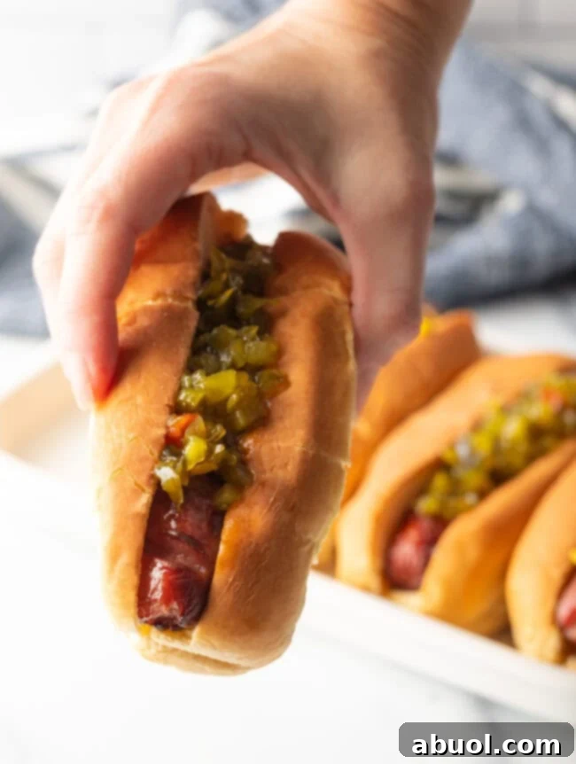 A hand holds a perfectly air-fried hot dog in a toasted bun, generously dressed with vibrant green relish, presented towards the camera.