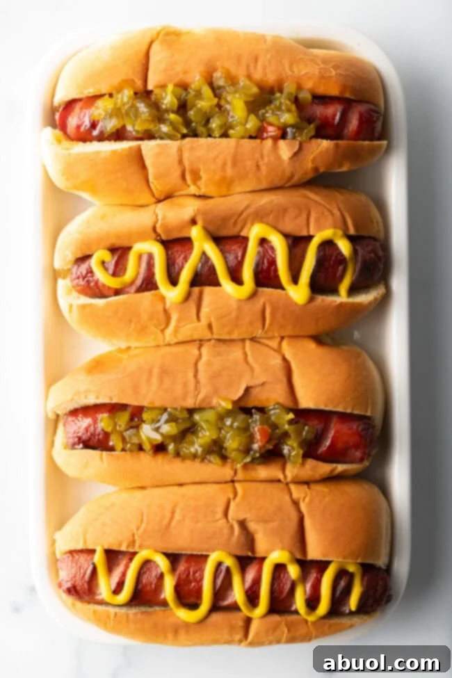A top-down view showcases four hot dogs in toasted buns, arranged side-by-side on a white rectangular plate. The toppings alternate between vibrant green relish and a classic zigzag of yellow mustard.