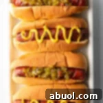 Top down view. Four hotdogs in buns placed side by side on a white rectangle plate. The toppings alternative between relish and zigzag of mustard.