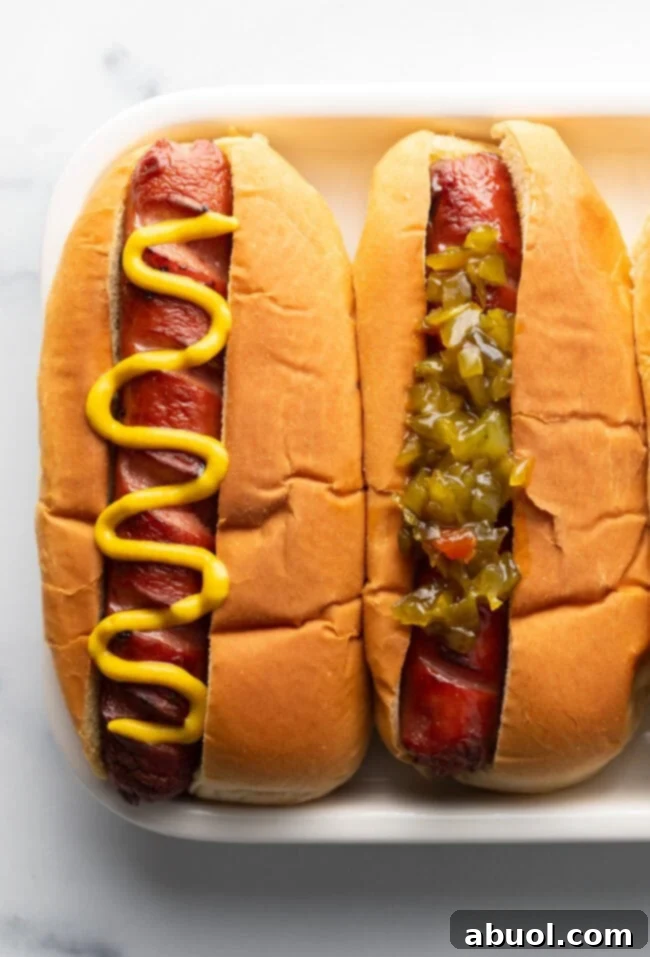 A top-down perspective showing two air-fried hot dogs on a white plate. One is adorned with a classic zigzag of yellow mustard, while the other is topped with vibrant green relish.
