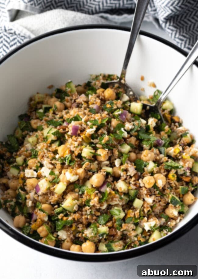 Large white bowl filled with bulgar wheat, veggies, and feta salad. Two metal salad spoons are in the bowl as well.