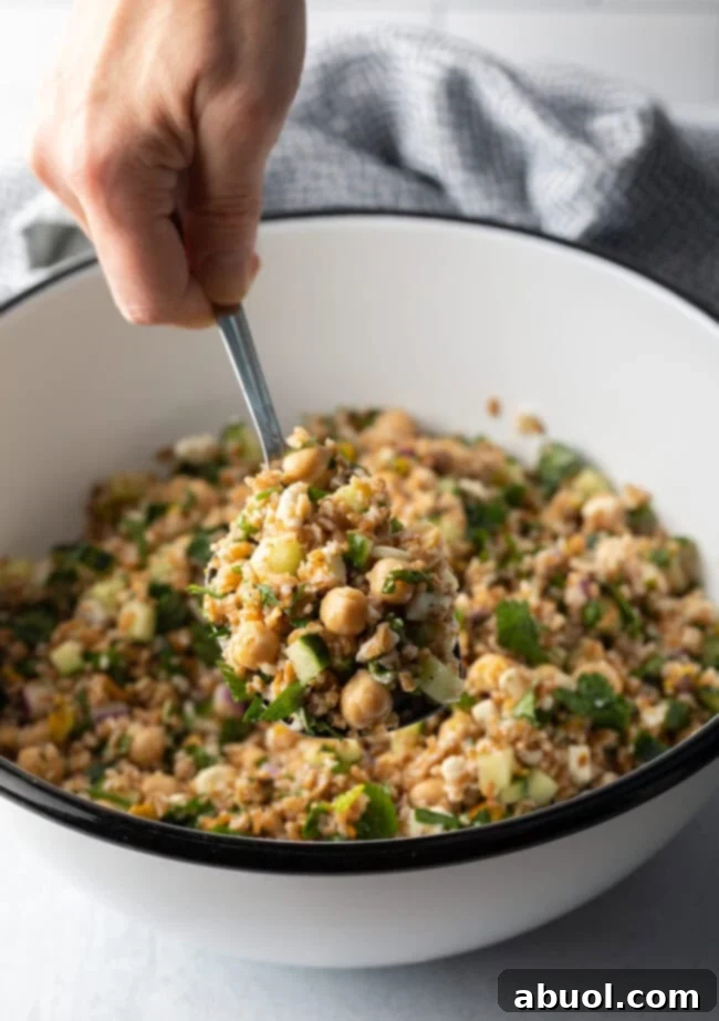 Hand with metal spoon scooping a serving of salad from a white bowl filled with bulgar wheat, veggies, and feta salad.