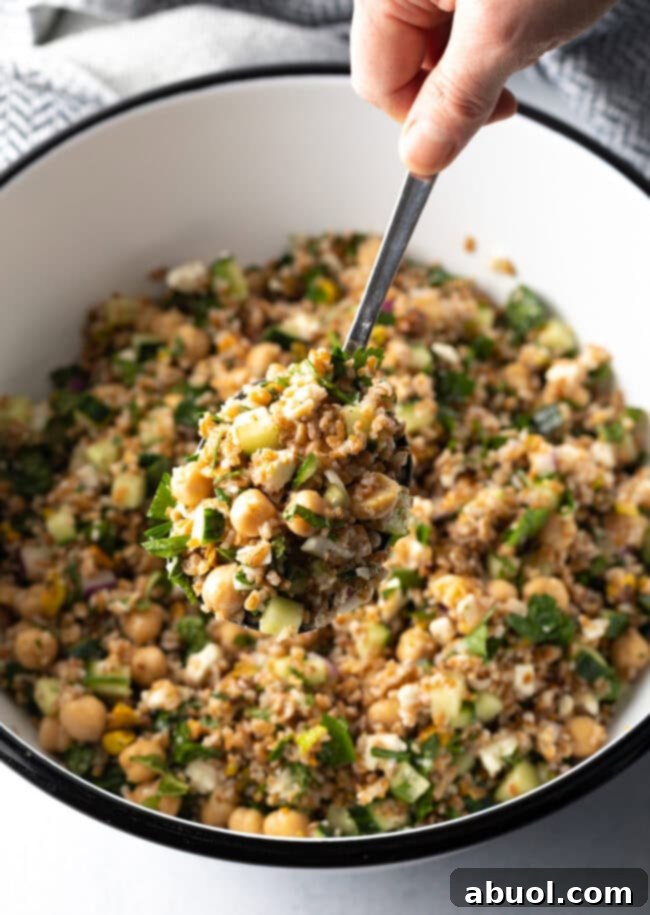 Hand with metal spoon scooping a serving of salad from a white bowl filled with bulgar wheat, veggies, and feta salad.