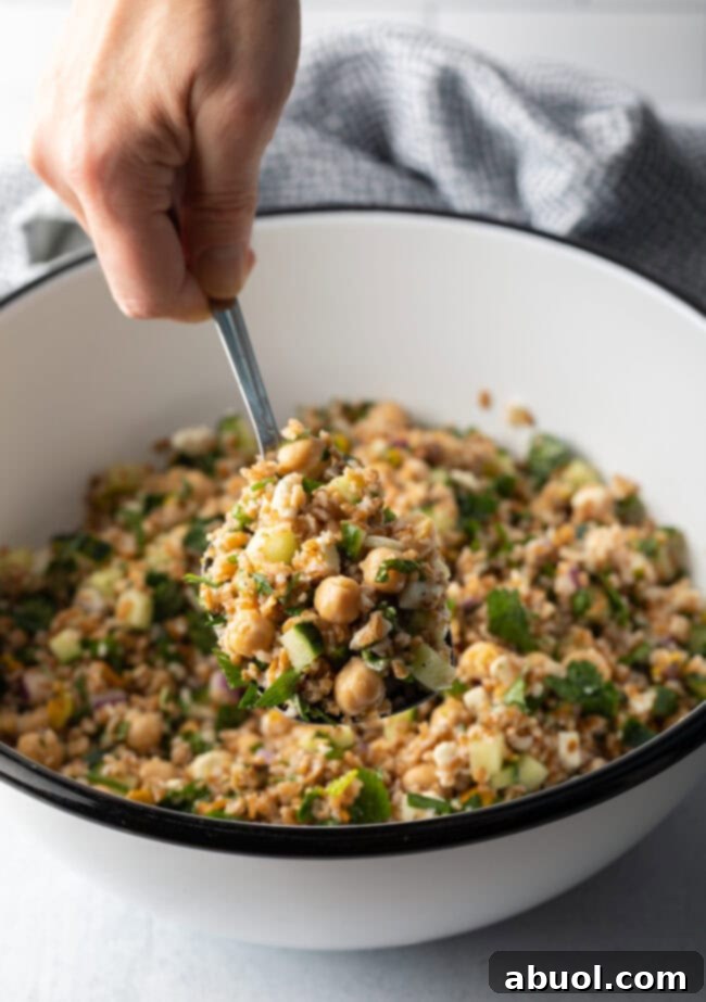 Hand with metal spoon scooping a serving of salad from a white bowl filled with bulgar wheat, veggies, and feta salad.