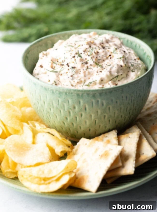 A vibrant green bowl brimmed with creamy clam dip, artfully placed on a crisp white plate and surrounded by an assortment of crunchy potato chips and savory crackers, ready for serving.