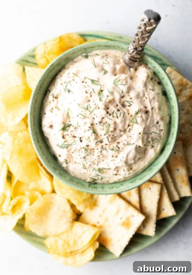 A beautiful top-down perspective of a vibrant green bowl, filled with our signature creamy clam dip, elegantly presented on a white plate and artfully surrounded by an inviting array of crispy potato chips and crunchy crackers.