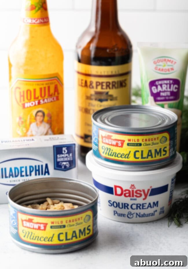 A carefully arranged collection of all the ingredients needed for the clam dip recipe, presented in their individual packaging. Featured items include a bottle of Cholula hot sauce, Worcestershire sauce, two cans of clams, a block of cream cheese, and a container of sour cream, all ready for preparation.