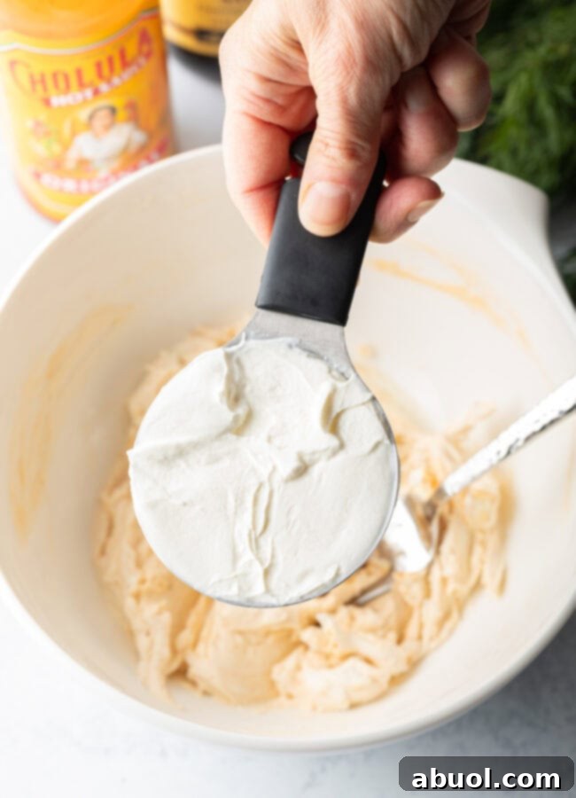 A hand holds a measuring cup, pouring sour cream into a white mixing bowl already containing seasoned cream cheese, illustrating a step in the clam dip preparation.