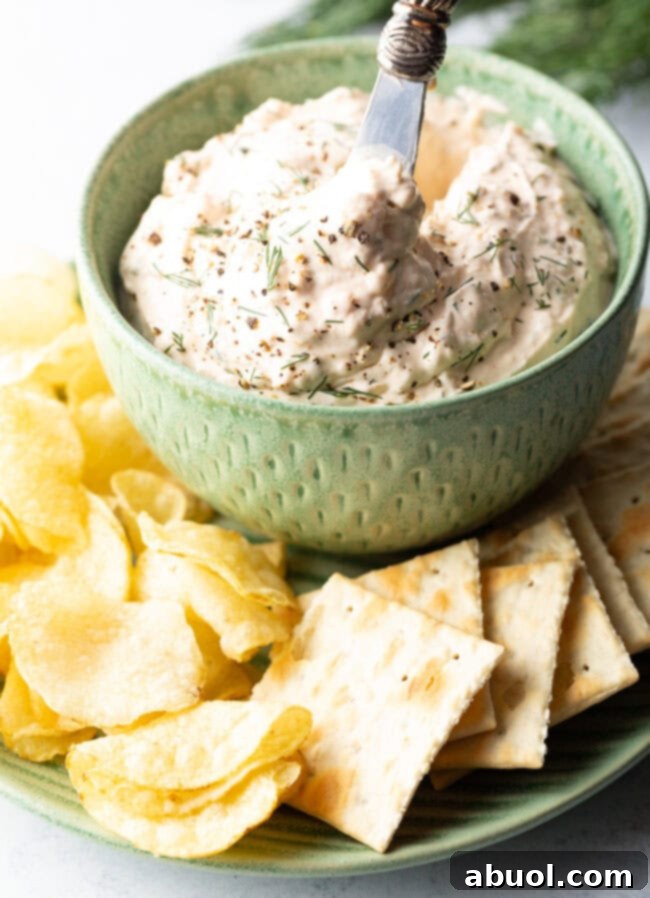 A green bowl brimming with creamy clam dip, beautifully positioned on a white plate, and encircled by a generous arrangement of crunchy potato chips and crisp crackers.