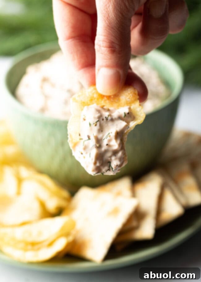 A hand gracefully holds a potato chip topped with a generous scoop of homemade clam dip, presented invitingly towards the camera for a close-up.