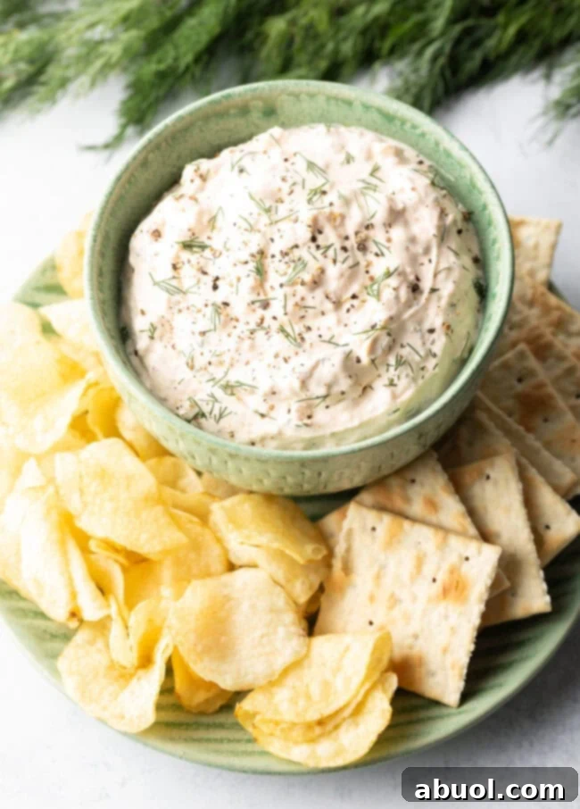 A vibrant top-down shot of a green bowl, generously filled with the best clam dip recipe, elegantly arranged on a white plate and surrounded by an inviting assortment of crispy potato chips and crunchy crackers.