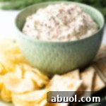 Green bowl filled with creamy clam dip, set on a white plate surrounded by potato chips and crackers.