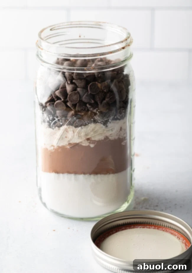 A large mason jar, open and filled with beautifully layered dry ingredients for homemade Ghirardelli brownie mix, positioned next to its lid, ready for assembly or gifting.