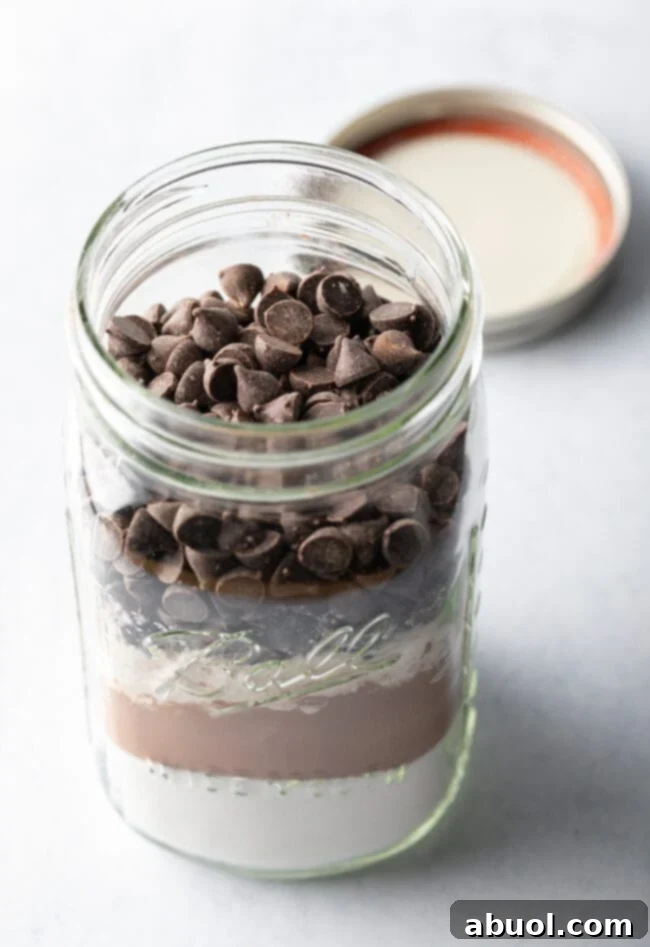 A close-up shot of a hand topping a clear glass jar of homemade brownie mix with extra Ghirardelli chocolate chips, highlighting its visual appeal for gifting.