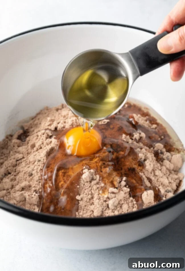 A hand adding vegetable oil from a small measuring cup into a white mixing bowl containing dry brownie mix and an egg, illustrating the wet ingredients step in brownie preparation.