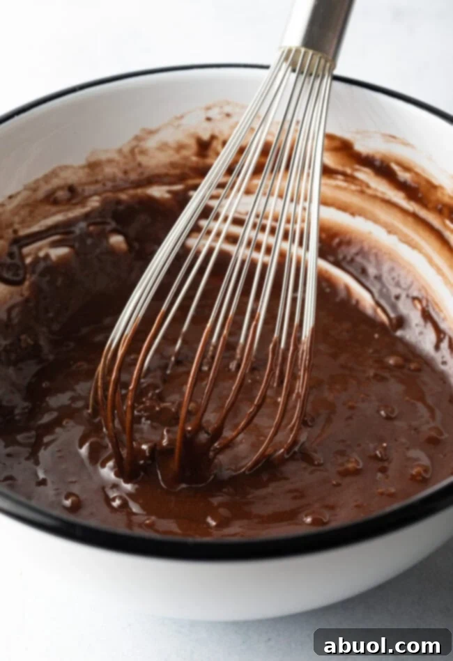 A metal whisk expertly combining wet and dry ingredients in a mixing bowl, transforming them into a rich, dark chocolate brownie batter, indicating the mixing process.