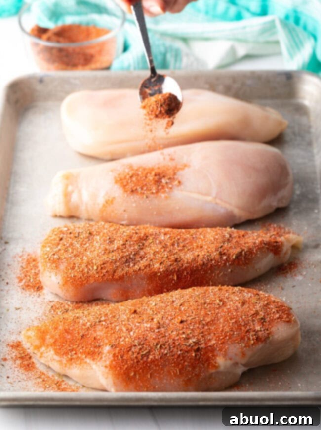 A metal spoon delicately sprinkles a rich, dark Cajun spice blend over raw chicken breasts, preparing them for the grill.
