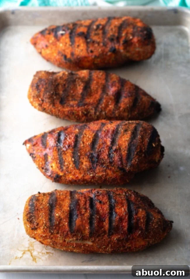 Four large, succulent grilled chicken breasts, boasting a rich blackened crust and infused with flavorful spices, presented on a serving tray.