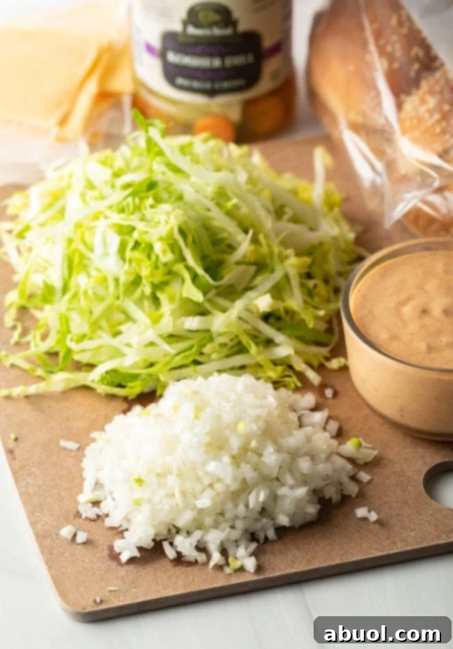 Cutting board with diced white onion, shredded iceberg lettuce, and glass bowl of homemade big mac sauce.
