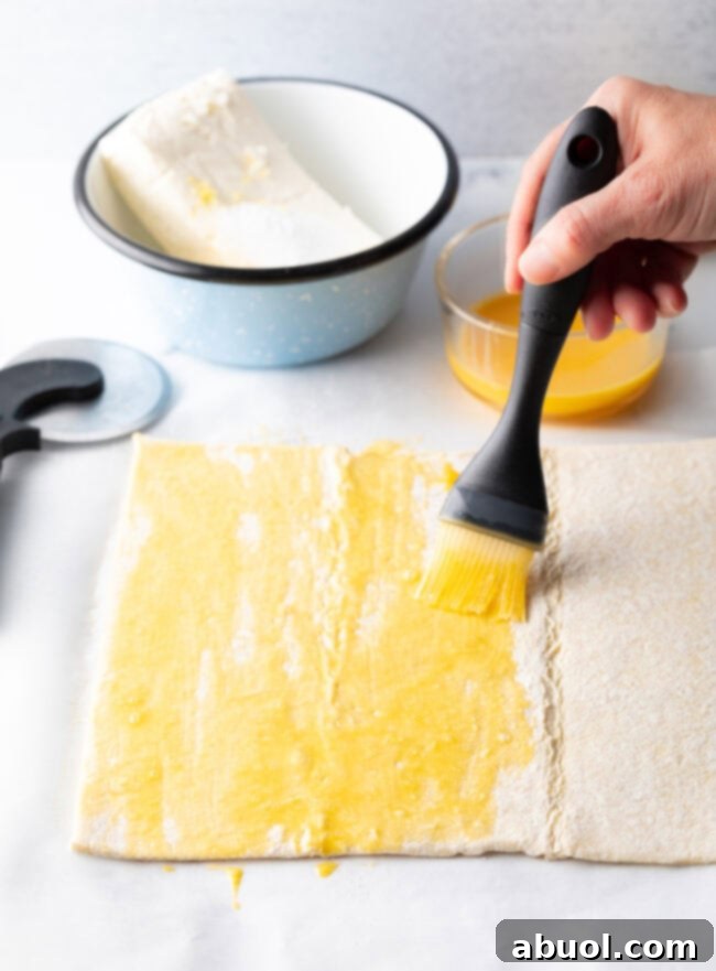Hand with pastry brush, applying egg wash on a sheet of puff pastry for a golden finish.